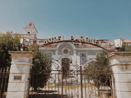 An old, ornate building with a sign reading 'Hotel Parque' at the entrance. The structure features decorative tiles, large windows, and a gated entrance with ironwork detailing. The building is surrounded by lush greenery, and the sky is clear.