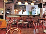 A cozy cafe or bar with several wooden chairs and tables arranged neatly. The room features a bar counter with various bottles displayed on shelves, and hanging pendant lights provide a warm ambiance. The walls are adorned with white subway tiles, and there is a television mounted above. A person is visible in the background near the bar.