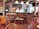A cozy cafe or bar with several wooden chairs and tables arranged neatly. The room features a bar counter with various bottles displayed on shelves, and hanging pendant lights provide a warm ambiance. The walls are adorned with white subway tiles, and there is a television mounted above. A person is visible in the background near the bar.