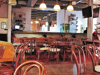 A cozy cafe or bar with several wooden chairs and tables arranged neatly. The room features a bar counter with various bottles displayed on shelves, and hanging pendant lights provide a warm ambiance. The walls are adorned with white subway tiles, and there is a television mounted above. A person is visible in the background near the bar.