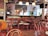 A cozy cafe or bar with several wooden chairs and tables arranged neatly. The room features a bar counter with various bottles displayed on shelves, and hanging pendant lights provide a warm ambiance. The walls are adorned with white subway tiles, and there is a television mounted above. A person is visible in the background near the bar.