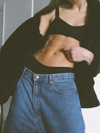 A person wearing a black top and loose blue jeans appears in the frame. Part of the person's torso is exposed, revealing defined abdominal muscles. The outfit includes a black jacket partially hanging off the shoulders.
