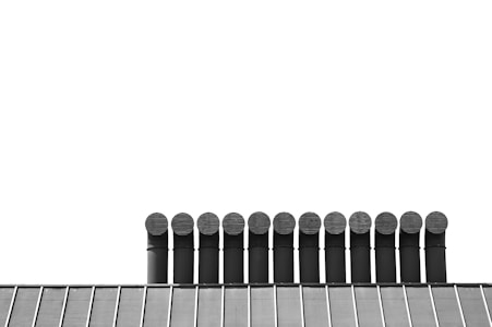 A row of industrial vents or exhaust pipes rise above a building's roof, which appears to be metallic and evenly segmented. The vents are uniformly shaped and closely aligned, set against a stark, clear white background.