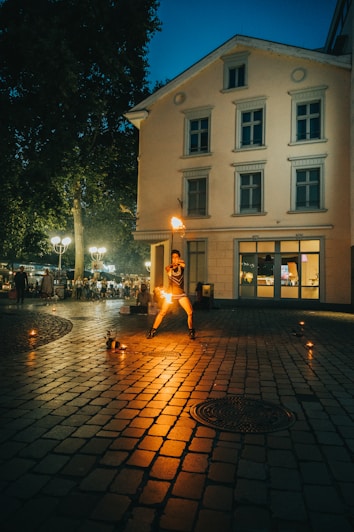 A performer juggles fire in a dimly lit cobblestone street in front of a multi-story building. A small audience can be seen in the background. The scene is illuminated by the warm glow of street lamps and the fire torches.