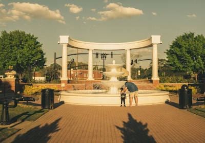 A person and a child stand in front of a large fountain in a park setting. The fountain is surrounded by white columns and greenery, with trees and a clear sky in the background. The brick pathway leads to the fountain, with benches and trash bins on the sides.