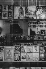 A display window with a variety of movie posters and DVD covers lined up on shelves. The collection includes both classic and contemporary films, and the setup has a vintage aesthetic due to the black and white photography. The titles and images are visible through the reflection of the glass, adding an artistic layer to the scene.