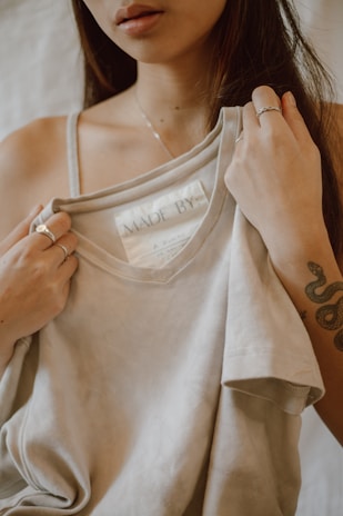 A woman is holding up a light beige t-shirt, revealing a tag inside that reads 'MADE BY:'. She is wearing several rings and has a floral tattoo on her arm. The background is softly blurred, giving the image an intimate and focused feel.