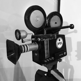 A vintage film camera with large reels and a lens protruding from the front. The camera features a logo marked 'OPEL' and various dials and buttons. It's positioned against a plain background, creating distinct shadows.