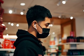 A person with short black hair is wearing a black mask and earbuds, browsing in a well-lit store with shelves of various products in the background.