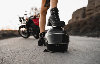 Sturdy riding boots with ankle protection standing on gravel beside a parked motorcycle.