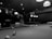 A black and white image depicts two people playing pool in a dimly lit pool hall. One person is holding a cue stick and leaning over the pool table, while the other appears to be interacting or assisting them. Several pool balls are scattered across the table, and the ceiling is adorned with hanging lights that illuminate the playing area.