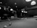 A black and white image depicts two people playing pool in a dimly lit pool hall. One person is holding a cue stick and leaning over the pool table, while the other appears to be interacting or assisting them. Several pool balls are scattered across the table, and the ceiling is adorned with hanging lights that illuminate the playing area.