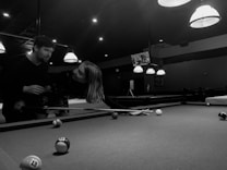 A black and white image depicts two people playing pool in a dimly lit pool hall. One person is holding a cue stick and leaning over the pool table, while the other appears to be interacting or assisting them. Several pool balls are scattered across the table, and the ceiling is adorned with hanging lights that illuminate the playing area.
