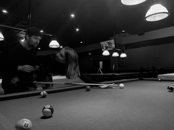 A black and white image depicts two people playing pool in a dimly lit pool hall. One person is holding a cue stick and leaning over the pool table, while the other appears to be interacting or assisting them. Several pool balls are scattered across the table, and the ceiling is adorned with hanging lights that illuminate the playing area.