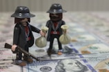 Two toy figures resembling outlaws or cowboys are holding money bags, one of which also holds a rifle. They are standing on a surface covered with hundred-dollar bills.
