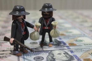 Two toy figures resembling outlaws or cowboys are holding money bags, one of which also holds a rifle. They are standing on a surface covered with hundred-dollar bills.