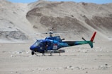 A blue and green helicopter with red accents is stationary on a barren, rocky desert terrain. The helicopter has some equipment attached to its underside. There are rugged, sandy hills in the background. One person is visible inside the helicopter.