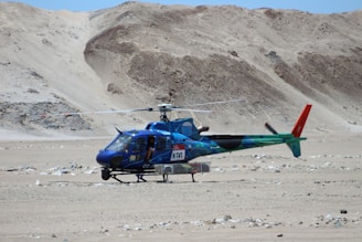 A blue and green helicopter with red accents is stationary on a barren, rocky desert terrain. The helicopter has some equipment attached to its underside. There are rugged, sandy hills in the background. One person is visible inside the helicopter.
