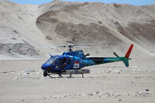 A blue and green helicopter with red accents is stationary on a barren, rocky desert terrain. The helicopter has some equipment attached to its underside. There are rugged, sandy hills in the background. One person is visible inside the helicopter.