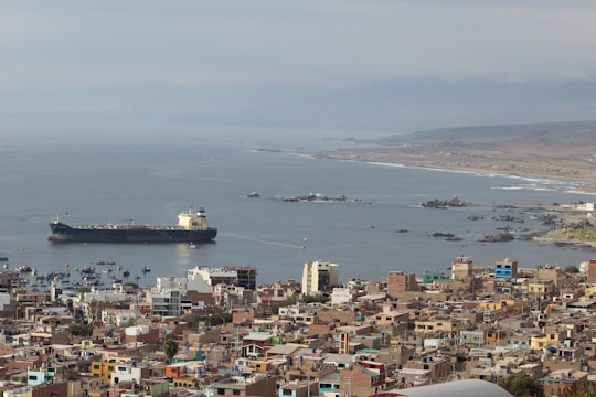 A large cargo ship is stationed in a coastal bay next to a densely built town. The shoreline is scattered with small fishing boats and rocky outcrops. The background displays a hazy horizon and distant hills under a cloudy sky.