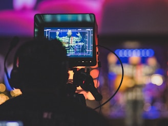 A person is sitting behind a large camera or video monitor, focusing on a screen displaying another person playing guitar. The scene is dimly lit with colorful, blurred lights in the background, suggesting a live performance or recording session.