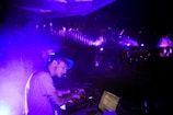 A DJ is focused on mixing music at a live event, with purple and blue lighting creating an electric atmosphere. The DJ is wearing a cap and using electronic equipment, while an audience is visible on an upper level, enjoying the performance.