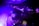 A DJ is focused on mixing music at a live event, with purple and blue lighting creating an electric atmosphere. The DJ is wearing a cap and using electronic equipment, while an audience is visible on an upper level, enjoying the performance.