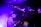 A DJ is focused on mixing music at a live event, with purple and blue lighting creating an electric atmosphere. The DJ is wearing a cap and using electronic equipment, while an audience is visible on an upper level, enjoying the performance.
