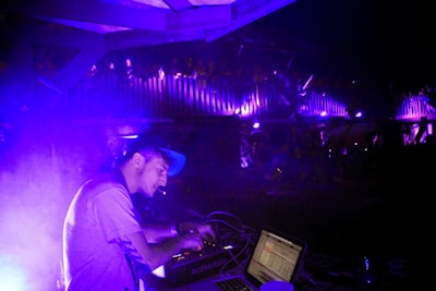 A DJ is focused on mixing music at a live event, with purple and blue lighting creating an electric atmosphere. The DJ is wearing a cap and using electronic equipment, while an audience is visible on an upper level, enjoying the performance.