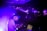 A DJ is focused on mixing music at a live event, with purple and blue lighting creating an electric atmosphere. The DJ is wearing a cap and using electronic equipment, while an audience is visible on an upper level, enjoying the performance.