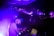 A DJ is focused on mixing music at a live event, with purple and blue lighting creating an electric atmosphere. The DJ is wearing a cap and using electronic equipment, while an audience is visible on an upper level, enjoying the performance.