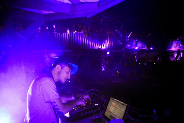 A DJ is focused on mixing music at a live event, with purple and blue lighting creating an electric atmosphere. The DJ is wearing a cap and using electronic equipment, while an audience is visible on an upper level, enjoying the performance.
