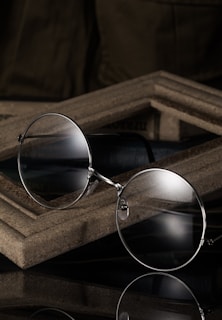 A pair of round eyeglasses with thin metallic frames is resting against a wooden, textured surface. The glasses reflect light subtly, suggesting a polished finish. The background is dark, creating a contrast that highlights the glasses.