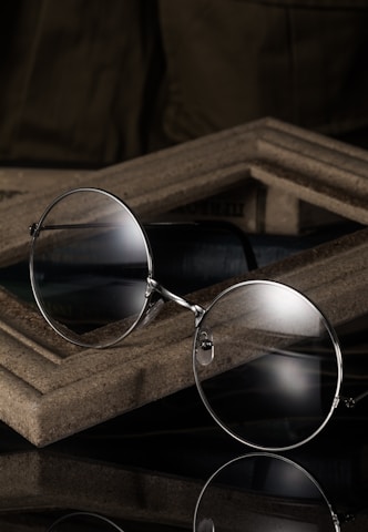 A pair of round eyeglasses with thin metallic frames is resting against a wooden, textured surface. The glasses reflect light subtly, suggesting a polished finish. The background is dark, creating a contrast that highlights the glasses.