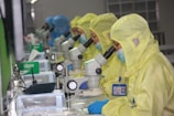 Several people in yellow protective suits and masks are working in a laboratory setting, each focused on looking through microscopes. The environment appears clean and organized, with multiple scientific tools and equipment on the desks.