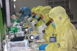 Several people in yellow protective suits and masks are working in a laboratory setting, each focused on looking through microscopes. The environment appears clean and organized, with multiple scientific tools and equipment on the desks.
