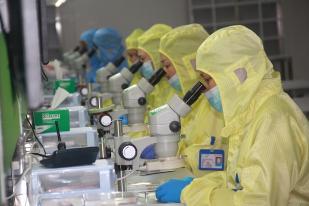 Several people in yellow protective suits and masks are working in a laboratory setting, each focused on looking through microscopes. The environment appears clean and organized, with multiple scientific tools and equipment on the desks.