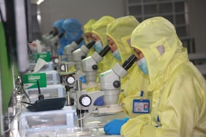 Several people in yellow protective suits and masks are working in a laboratory setting, each focused on looking through microscopes. The environment appears clean and organized, with multiple scientific tools and equipment on the desks.