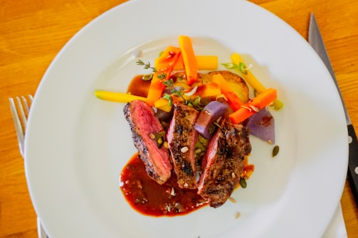 A neatly plated dish with slices of cooked meat accompanied by vibrant vegetables including carrots, potatoes, and purple onions. The meat is garnished with herbs and seeds, all drizzled with a rich sauce. The setting includes a white plate on a wooden surface with a knife and fork placed nearby.