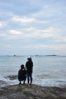 Two people stand on a rocky shoreline, facing the ocean under a cloudy sky. One person is squatting while the other is standing beside them, gently holding their shoulder. The scene conveys a sense of companionship and tranquility as the waves lightly crash against the rocks.