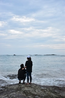 Two people stand on a rocky shoreline, facing the ocean under a cloudy sky. One person is squatting while the other is standing beside them, gently holding their shoulder. The scene conveys a sense of companionship and tranquility as the waves lightly crash against the rocks.
