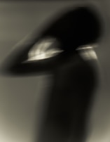 A blurred silhouette of a person seems to be clasping their head with one hand in a dimly lit environment. The contours are indistinct, creating an abstract impression of motion or uncertainty.