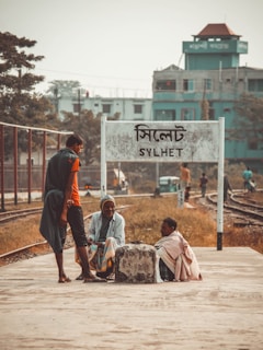 Three people are gathered at a railway platform, one standing and two sitting near a concrete block. They appear to be engaged in conversation with a background of railway tracks and a sign that reads 'SYLHET'. A few buildings are visible in the distance with muted greenery surrounding the area.