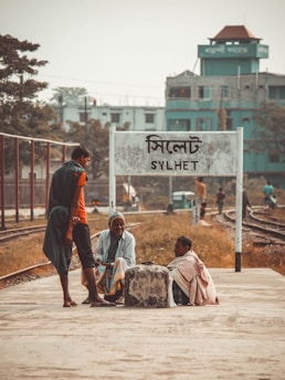 Three people are gathered at a railway platform, one standing and two sitting near a concrete block. They appear to be engaged in conversation with a background of railway tracks and a sign that reads 'SYLHET'. A few buildings are visible in the distance with muted greenery surrounding the area.