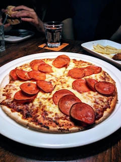 A pepperoni pizza is placed on a white plate, resting on a wooden table. The pizza is topped with sliced pepperoni and melted cheese. In the background, there is a glass of water and a person holding a sandwich. A side dish of fries is also partially visible.