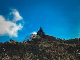 A rustic church with a cross on top sits atop a hill covered in lush vegetation. The sky is expansive and bright blue, dotted with white clouds.