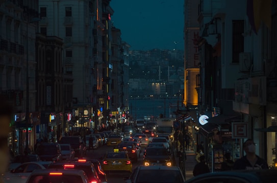 A bustling city street at dusk, framed by tall buildings on both sides. The street is filled with heavy traffic, featuring numerous cars and taxis. Bright neon lights from shops and signs illuminate the scene, while a view of a distant waterway and hills is visible in the background under a darkening sky.