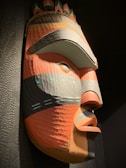 A large, colorful tribal mask carved from wood with prominent geometric patterns. The mask features bold colors such as red, orange, gray, and black, and has distinct facial features with exaggerated eyes and lips. Small circular decorations are placed above the eyes and around the cheeks, adding to the intricacy of the design.