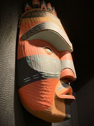A large, colorful tribal mask carved from wood with prominent geometric patterns. The mask features bold colors such as red, orange, gray, and black, and has distinct facial features with exaggerated eyes and lips. Small circular decorations are placed above the eyes and around the cheeks, adding to the intricacy of the design.
