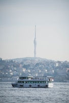A ferry is sailing in a body of water with a cityscape in the background. A tall, modern telecommunications tower is visible above a densely built hillside, conveying a mix of urban and natural landscapes.